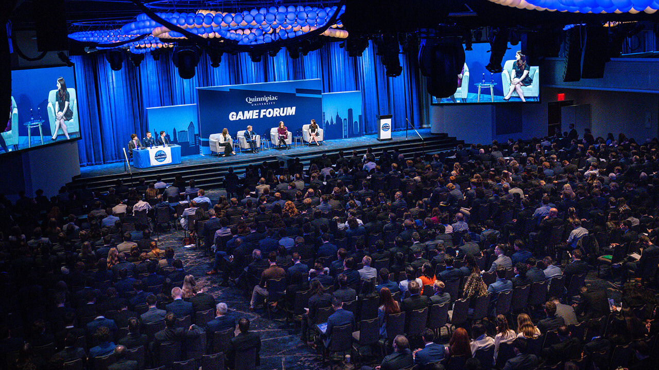 A large auditorium with every seat filled while a panel speaks on a stage with a GAME Forum sign behind them