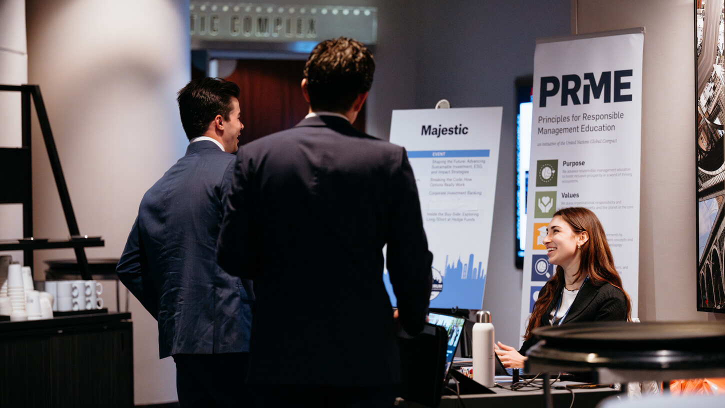 A representative at the PRME booth smiles while engaging with students near a sign promoting sustainable leadership values.