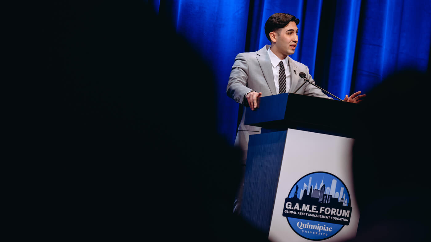A student in a light gray suit speaks at the podium during the Quinnipiac GAME Forum, addressing the audience against a blue curtain backdrop.