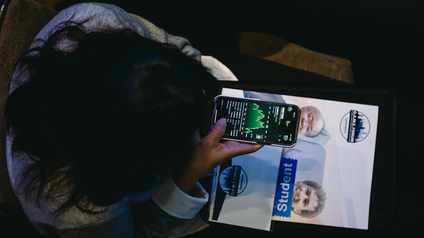 An overhead view of a student checking market data on their phone during the forum, with a GAME Forum program in hand.