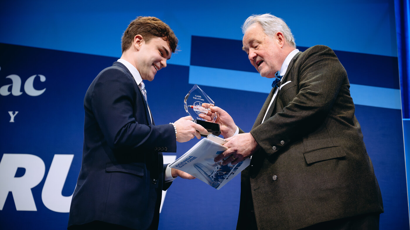 A student awards Tom Keene with a trophy on stage, inducting him into the GAME Hall of Fame.
