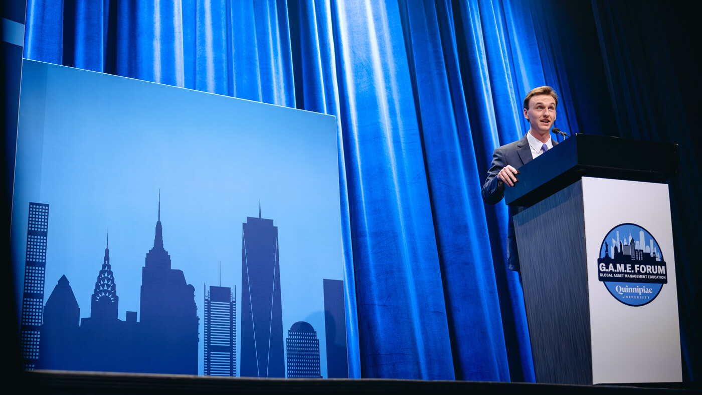 A student stands at the GAME Forum podium, presenting in front of a cityscape backdrop.