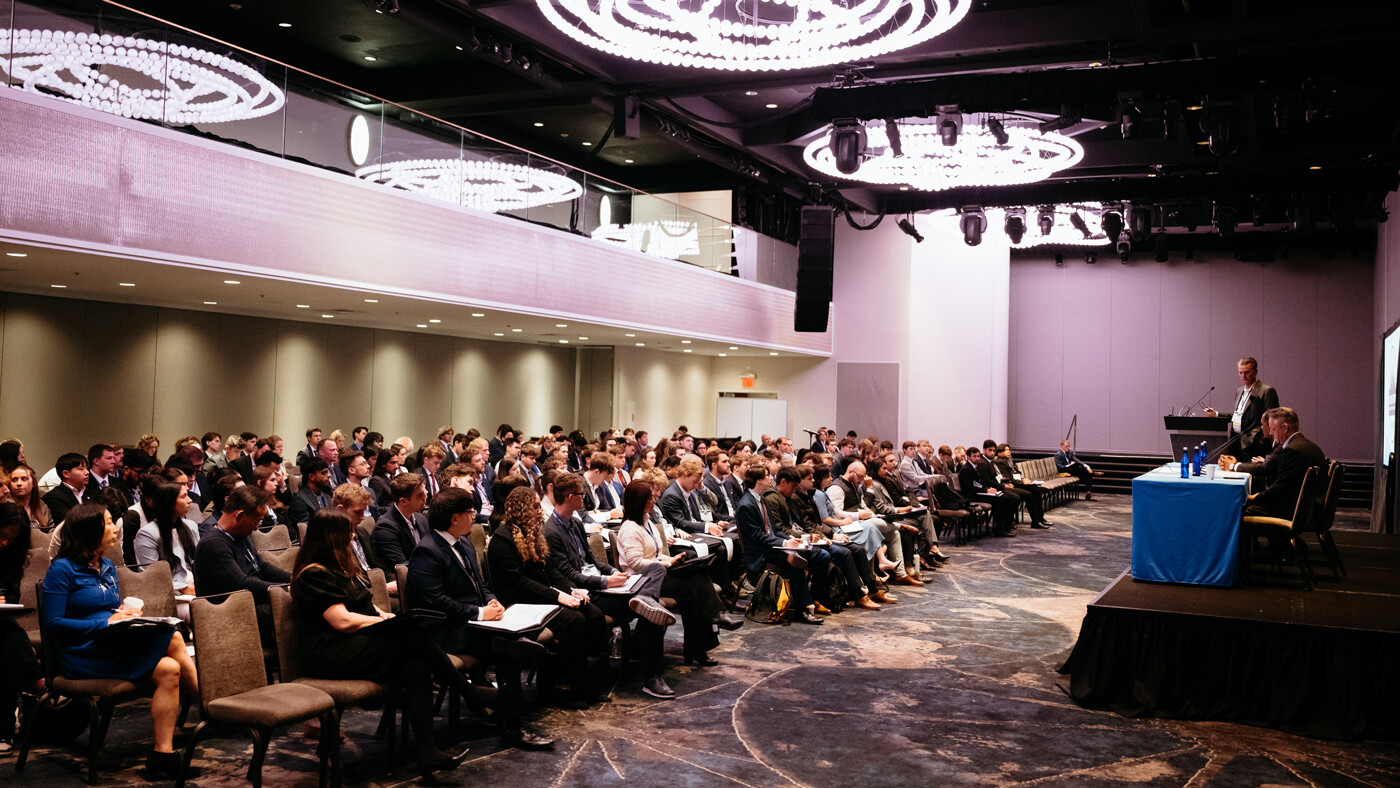 A speaker stands at a podium while a panelist listens onstage; the audience fills the room during a GAME Forum keynote presentation.