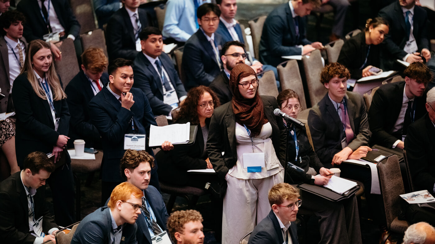 A student stands at a microphone to ask a question during a Q&A session, with peers seated around them observing.