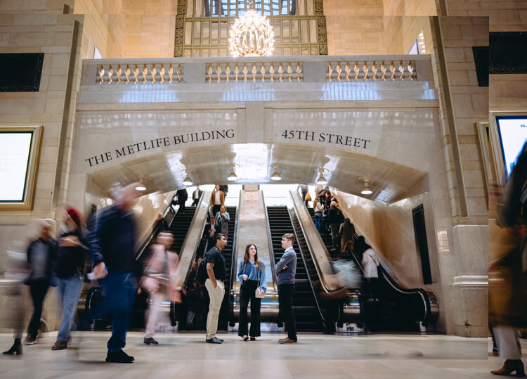 Three Quinnipiac students stand talking at the base of escalators inside Grand Central Terminal as commuters blur past in motion.