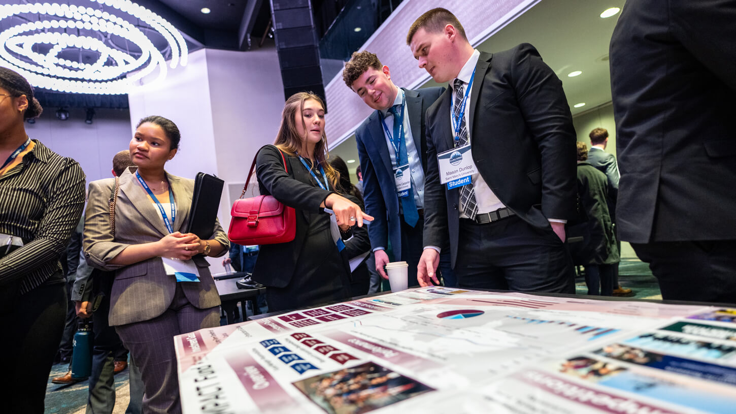 A female student points to a section of a flat poster display while surrounded by engaged peers from various universities.