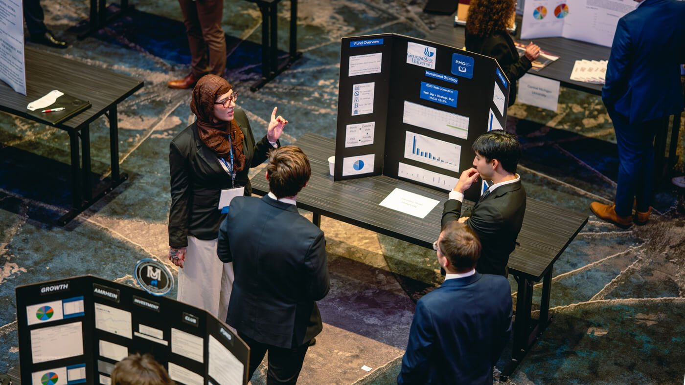 A student presenter from Georgia State University discusses their investment fund strategy with several attendees, standing beside a poster with bar charts and pie graphs.