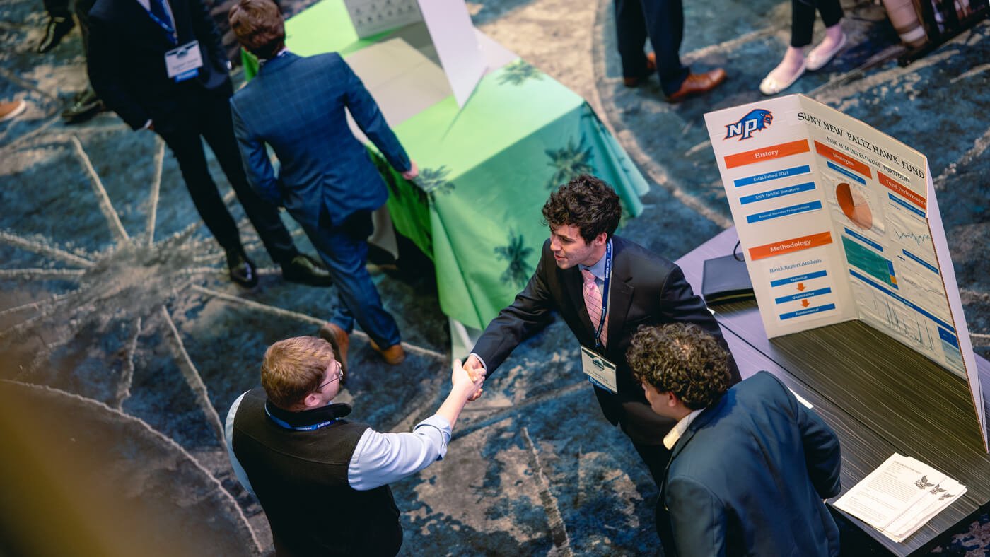 A student representative from SUNY New Paltz shakes hands with a visitor next to a colorful tri-fold poster labeled “Hawk Fund” highlighting their fund’s methodology and history.