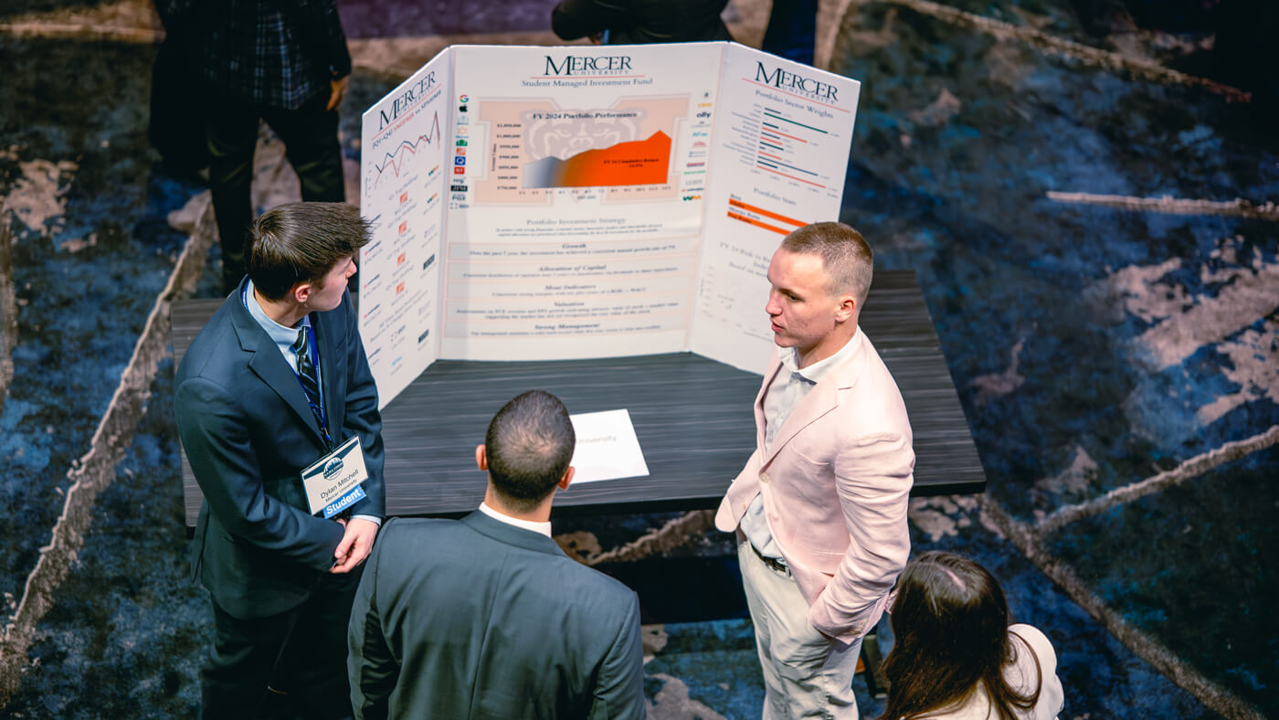 A student from Mercer University explains a detailed financial poster to a small group. The display includes portfolio performance graphs and sector weightings.