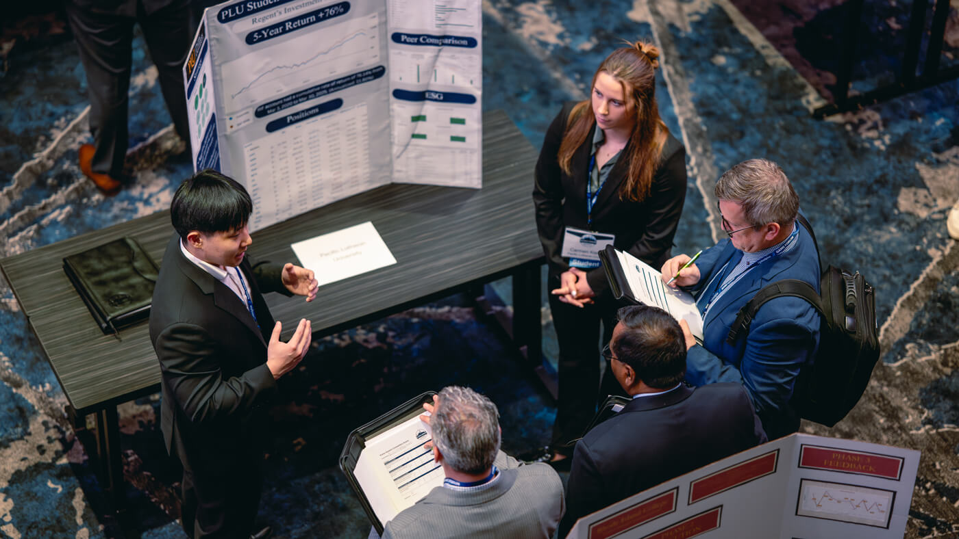 A student from Pacific Lutheran University presents a financial portfolio to a group of judges at the GAME Forum, using a poster that highlights a 5-year return of +76%.