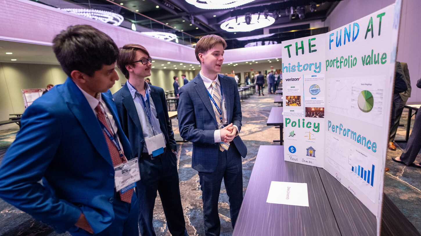 Three student presenters stand next to a poster board titled “The Fund at Drew University” that features charts, logos, and handwritten headings on portfolio value, policy, and performance.
