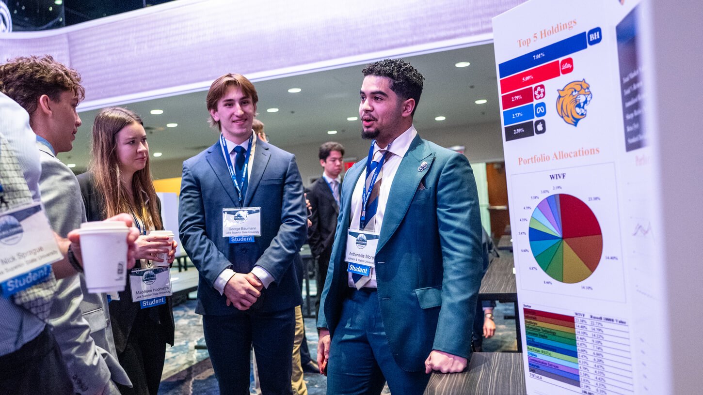 A student presenter from Johnson & Wales University explains a portfolio poster showing top stock holdings and a colorful allocation pie chart to a group of fellow students.