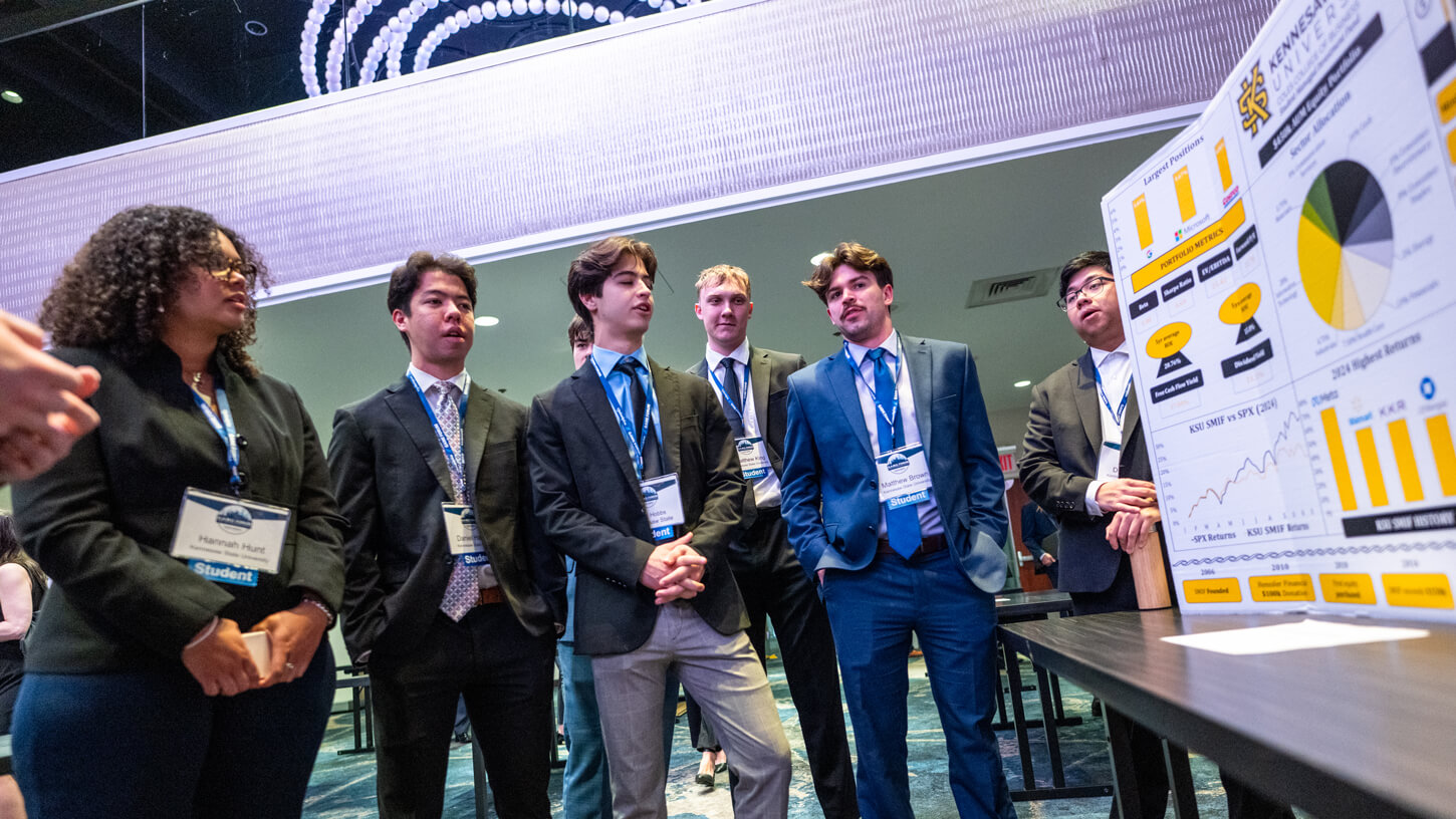 A team of students from Kennesaw State University attentively listens while one of them presents a poster with bold yellow graphs, pie charts, and return comparisons.